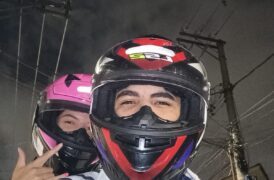 Homem com capacete vermelho e, atrás, uma mulher com capacete rosa. Ambos sorriem em cima de uma moto